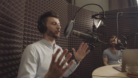 bearded young radio broadcaster explains something to a person sitting next to him in a radio recording studio