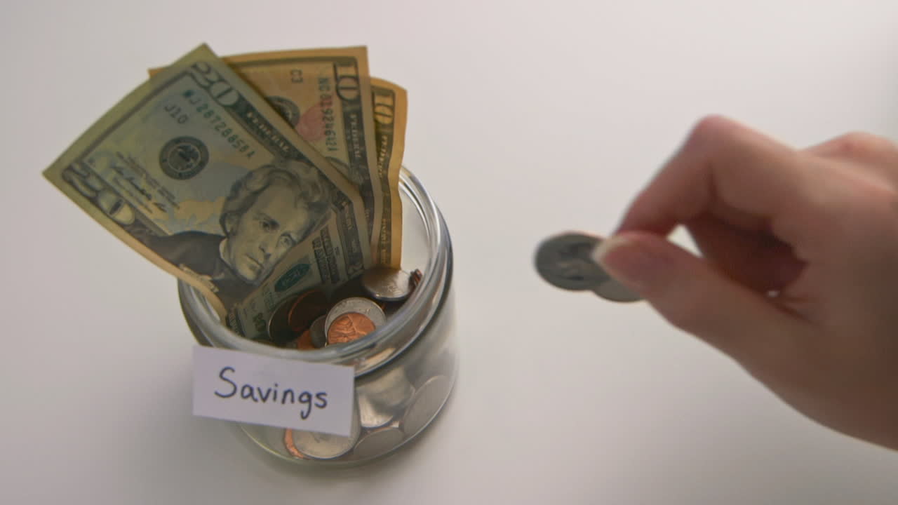 A caucasian woman's hand puts two American quarters into a &amp;quot;Savings&amp;quot; jar
