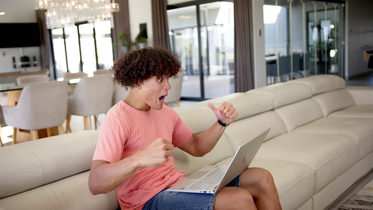 Excited man sitting on couch, using laptop and celebrating success at home