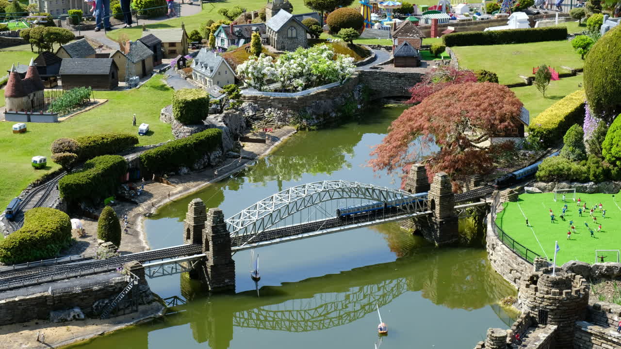 Extreme Wide Shot of Miniature Model Village in Traditional English Countryside Featuring River with Sailing Boats, Bridge, Old Locomotive, Church, Castle, Football Field, and Fun Fair in the Distance