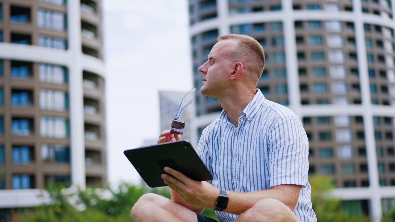 Man viewing a tablet with glasses held near his mouth. He looks down at the screen in a striped shirt, with curved buildings blurred in the background.