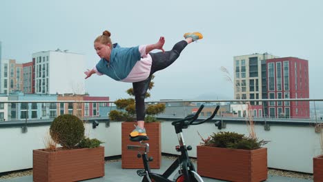 athletic girl performing swallow acrobatic trick exercises on cycling stationary bike on house roof