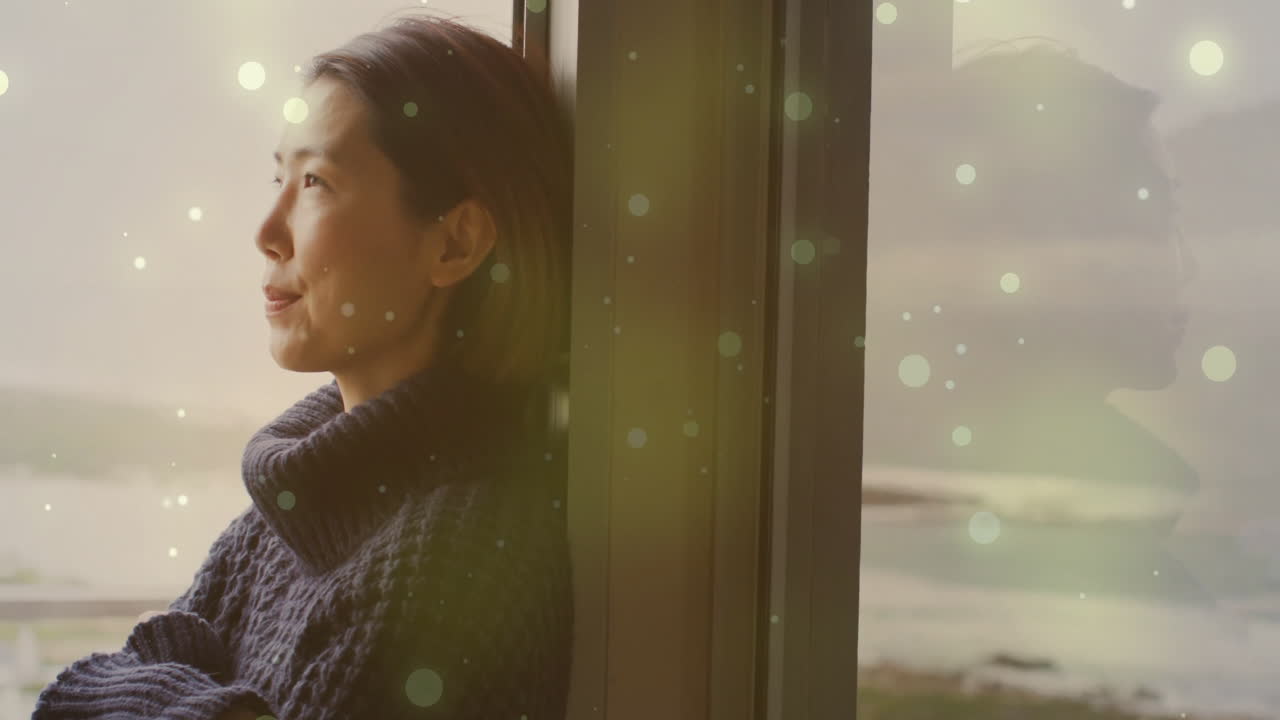 Animation of light spots over asian woman looking through window at home