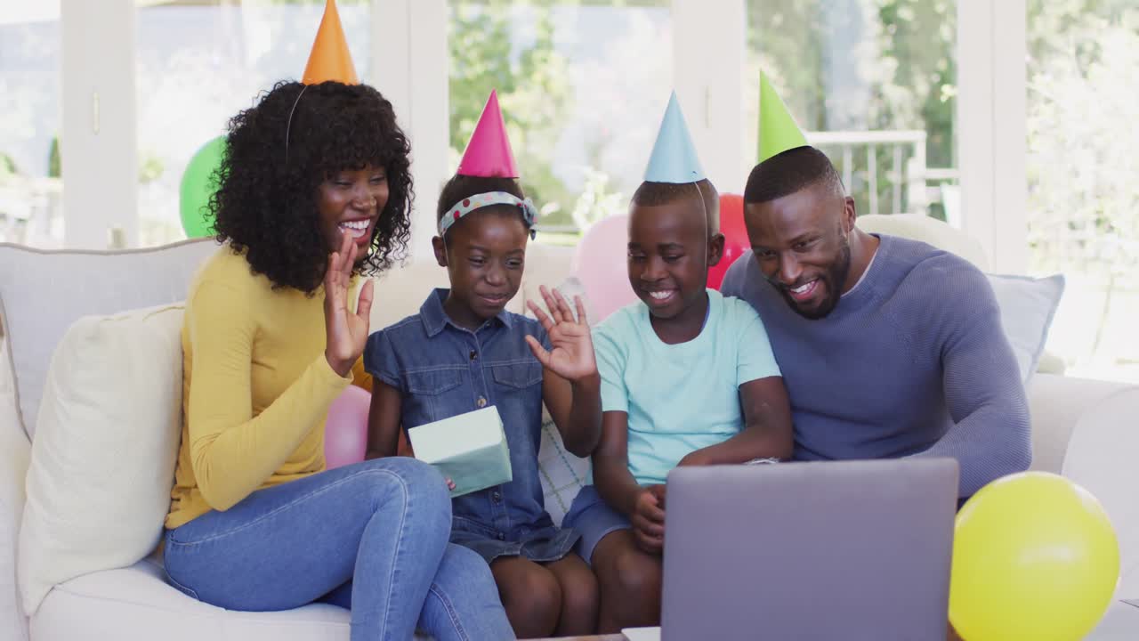 African american family in party hats waving having a videocall on laptop while sitting on the couch
