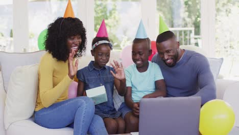 Familia-Afroamericana-Con-Sombreros-De-Fiesta-Saludando-Con-Una-Videollamada-En-Una-Computadora-Portátil-Mientras-Se-Sienta-En-El-Sofá