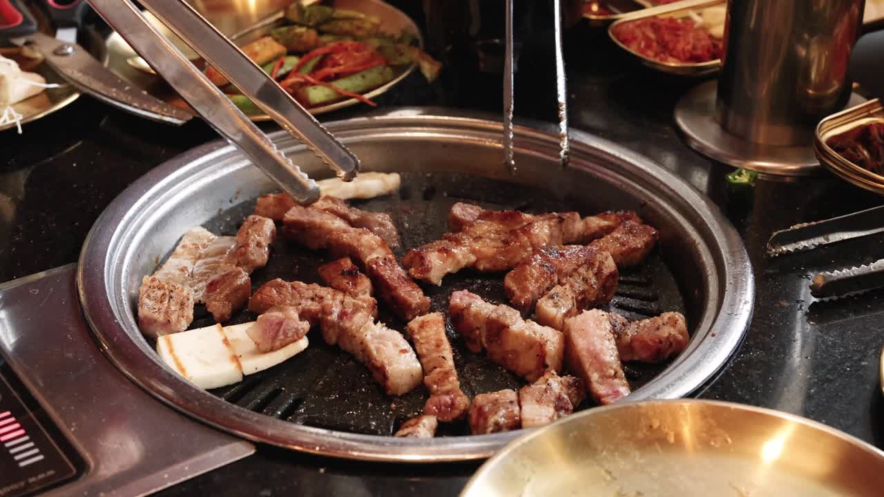 Hand uses tongs to flip pork belly pieces on tabletop grill, warm restaurant lighting