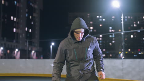 futbolista con capucha joggling fútbol durante la práctica nocturna en el campo deportivo urbano rodeado de edificios de gran altura iluminados y luces de calle brillantes