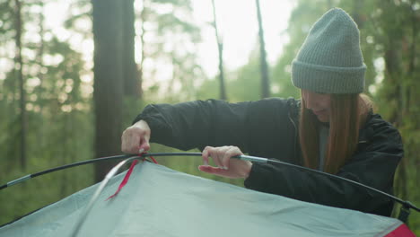 young woman wearing beanie gently smiles while loosening red rope from flexible tent pole in morning forest setting surrounded by trees and natural light as she prepares camp with calm focus