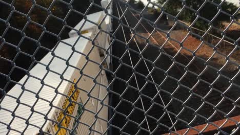 train passing beneath overpass behind a chainlink fence.