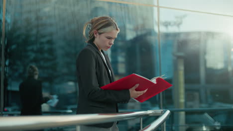 lady dressed in black blazer reading through red book thoughtfully near glass building as sunlight ambiance reflects surroundings on glass wall