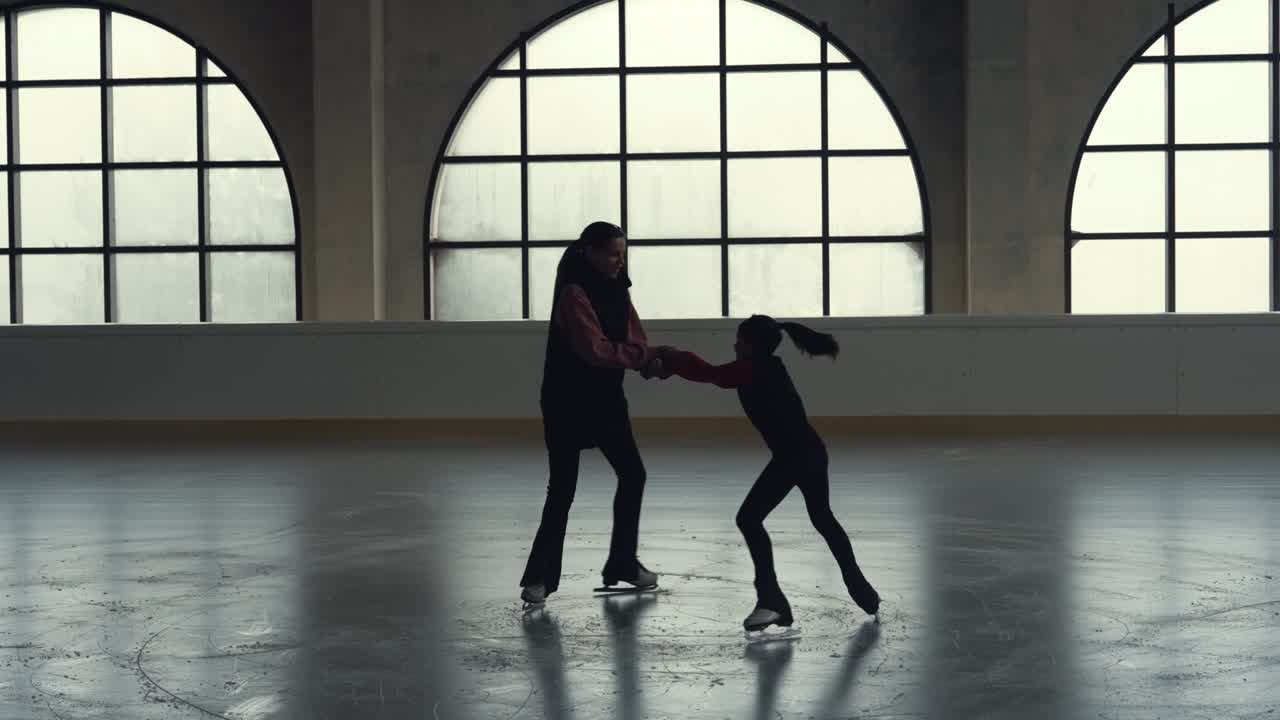 Figure skating training. Woman coach and her student girl are circling on ice arena holding hands. Little girl skater is skating with her trainer in dark stadium. Dark silhouettes. Slow motion