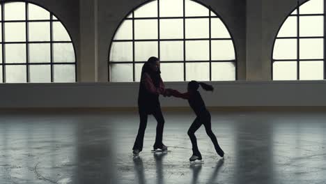 figure skating training. woman coach and her student girl are circling on ice arena holding hands. little girl skater is skating with her trainer in dark stadium. dark silhouettes. slow motion