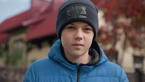 close-up of boy wearing black beanie and blue jacket outdoors, looking calm and thoughtful, with a blurred red-roofed house and vibrant autumn trees in background