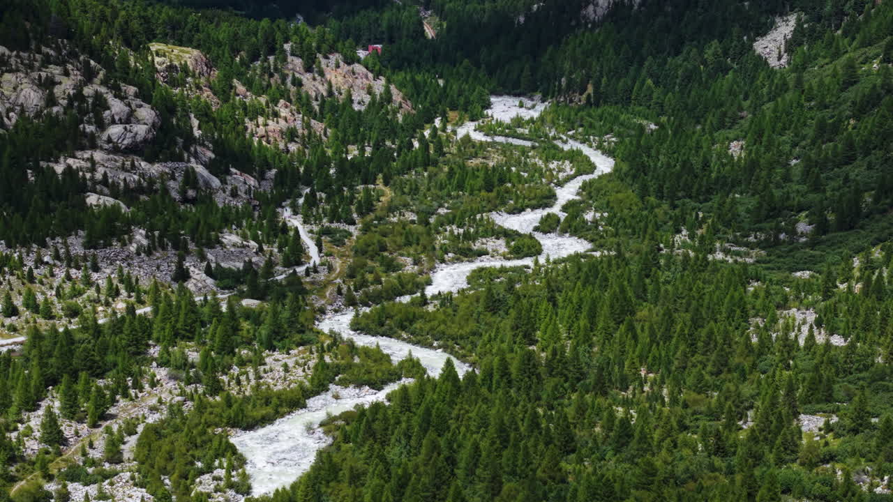 Aerial view pans above icy river meandering between pine trees and rocky mountain slopes
