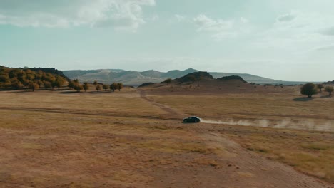 Tracking-drone-shot-of-a-black-car-driving-on-a-country-road-at-sunset,-dusty-road,-travel,-cinematic-grade