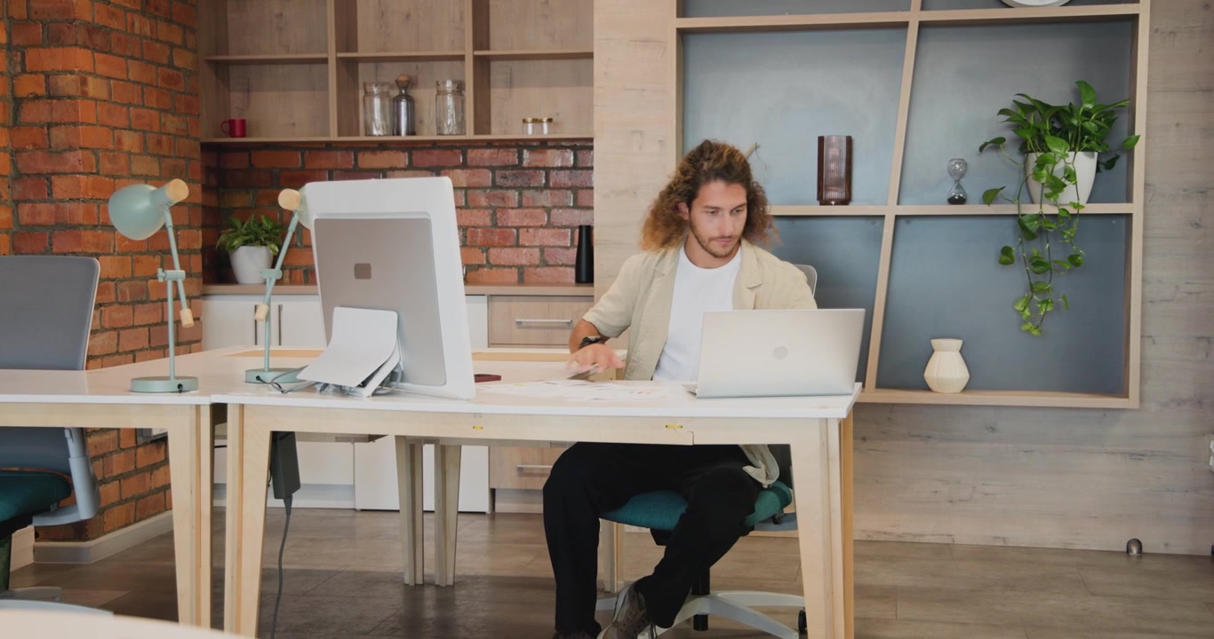 Uomo in camicia casual che parla al telefono, termina la chiamata, sistema dei documenti e digita al laptop sulla scrivania.