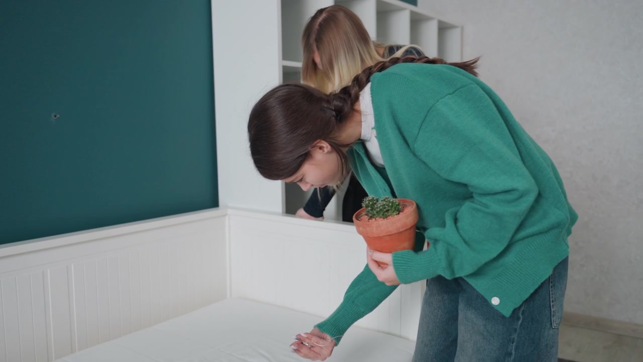 Young woman arranging plant on bed, carefully checks mattress surface and bed frame while companion watches, modern apartment, soft natural light, terracotta pot, cozy teal accent wall, interior