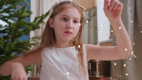 close-up of little girl in white gown with flower in her hair carefully arranging christmas lights for tree decoration with focus on her hands adjusting the lights during holiday preparation