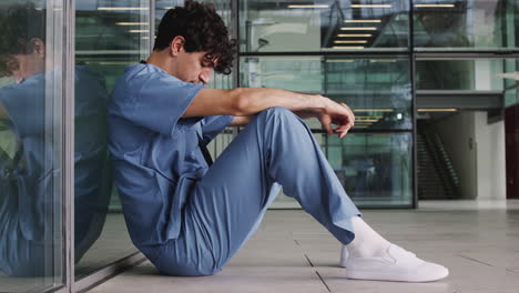 Cansado-Joven-Médico-Vistiendo-Batas-Sentado-Contra-La-Pared-En-El-Moderno-Edificio-Del-Hospital