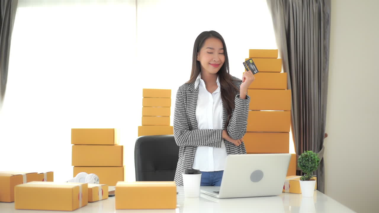 una joven y exitosa mujer de negocios rodeada de cajas que esperan ser enviadas sostiene su tarjeta de crédito en señal de triunfo