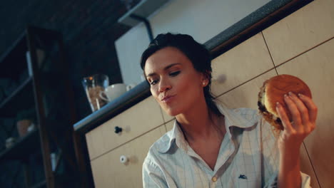 una mujer hambrienta comiendo una hamburguesa en la cocina de su casa. una chica sonriente bebiendo vino tinto.