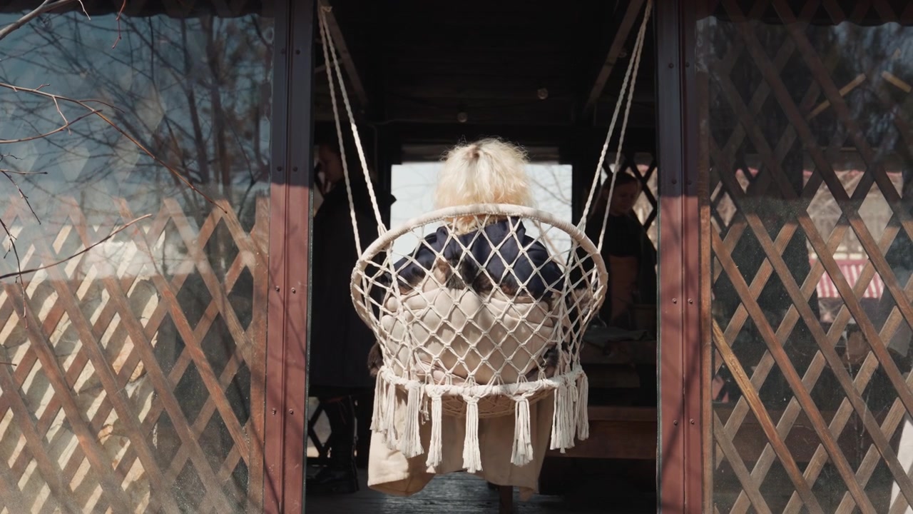 Young child seen from behind sits in woven macrame hanging chair at rustic porch doorway, gently swinging beside wooden floorboards as soft daylight highlights fringe, ropes, and cozy home details