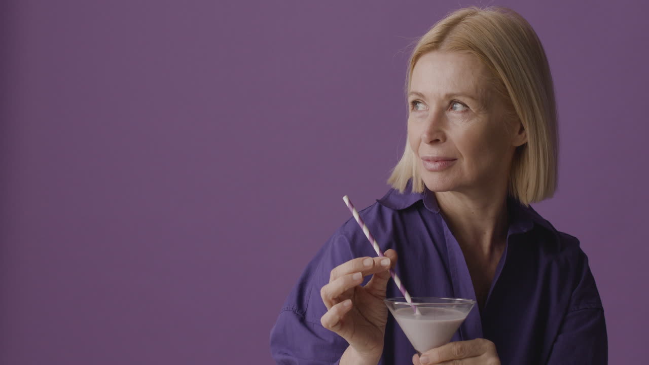 Blonde Mature Woman With Purple Shirt Posing Holding A Cocktail And Smiling At Camera On Purple Background