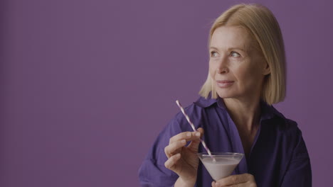 mujer madura rubia con camisa morada posando sosteniendo un cóctel y sonriendo a la cámara sobre fondo morado