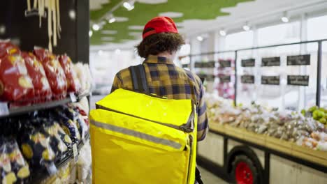 Rear-view-of-a-confident-brunette-guy-with-curly-hair-in-a-checkered-shirt-with-a-yellow-bag-a-food-delivery-man-walks-near-the-counter-in-a-supermarket-and-selects-the-necessary-products