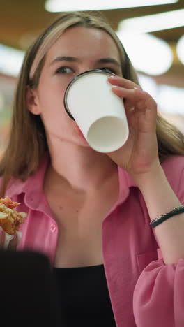 mujer de vestido rosa tomando un sorbo de café mientras sostiene una hamburguesa en su mano izquierda, sentada en un restaurante del centro comercial, continúa trabajando después de tomar una copa, con un fondo ocupado y borroso