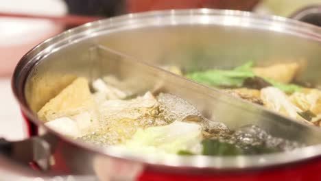 close-up of a divided hotpot simmering with fresh vegetables and bubbling broth in a red pot.