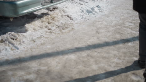 low angle leg view of people walking past parked car on icy sidewalk, winter sun casting long shadows over snow and slush, boots and sneakers stepping along gritty pavement during cold urban commute