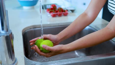hands thoroughly rinse green limes under running water in a modern kitchen sink with natural daylight and strawberries visible in the background