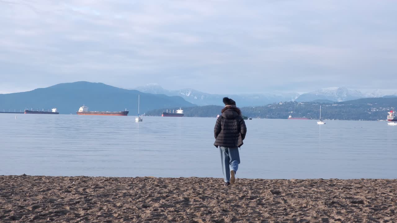 Young woman walking and skipping happily on a beach wearing a black beanie, sun glasses, black puffy coat and jeans on a beautiful winter day (4K)