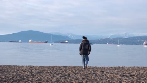 young woman walking and skipping happily on a beach wearing a black beanie, sun glasses, black puffy coat and jeans on a beautiful winter day (4k)