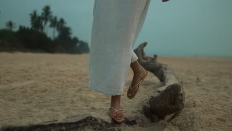 Woman-in-a-white-dress-walking-on-a-large-piece-of-driftwood-on-a-sandy-beach