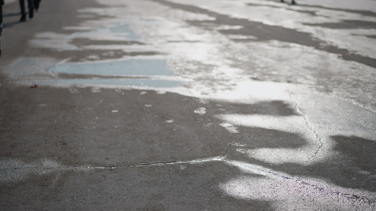 winter road surface with cracks and melting snow, bright sun reflection glistens across wet asphalt, thin mist and puddles sparkle, gritty texture and cold light suggest thaw on city pavement
