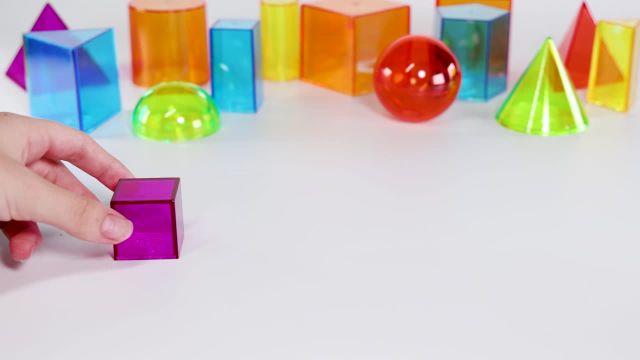 Hands manipulate vibrant geometric shapes on a white surface. Bright lighting highlights the colorful prisms, spheres, and cubes