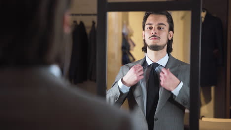 satisfied young man trying on suit in front of mirror in fashion store