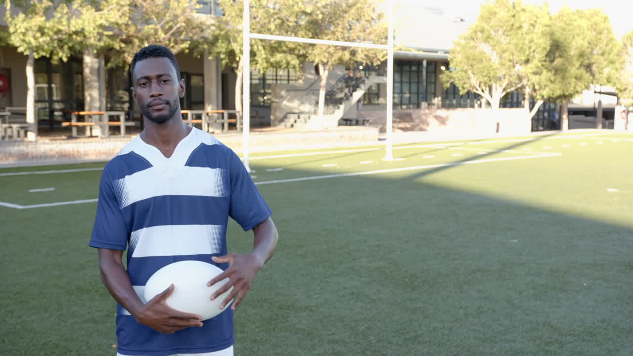 Holding rugby ball, african american man standing on sports field in striped jersey, copy space