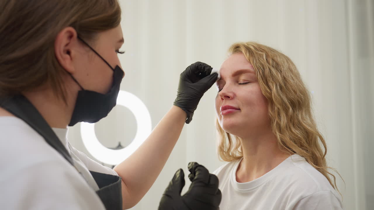 Beautician wearing black gloves gently wipes eyebrow area of blond client with cotton pad in well-lit beauty salon setting, preparing skin for treatment or makeup application