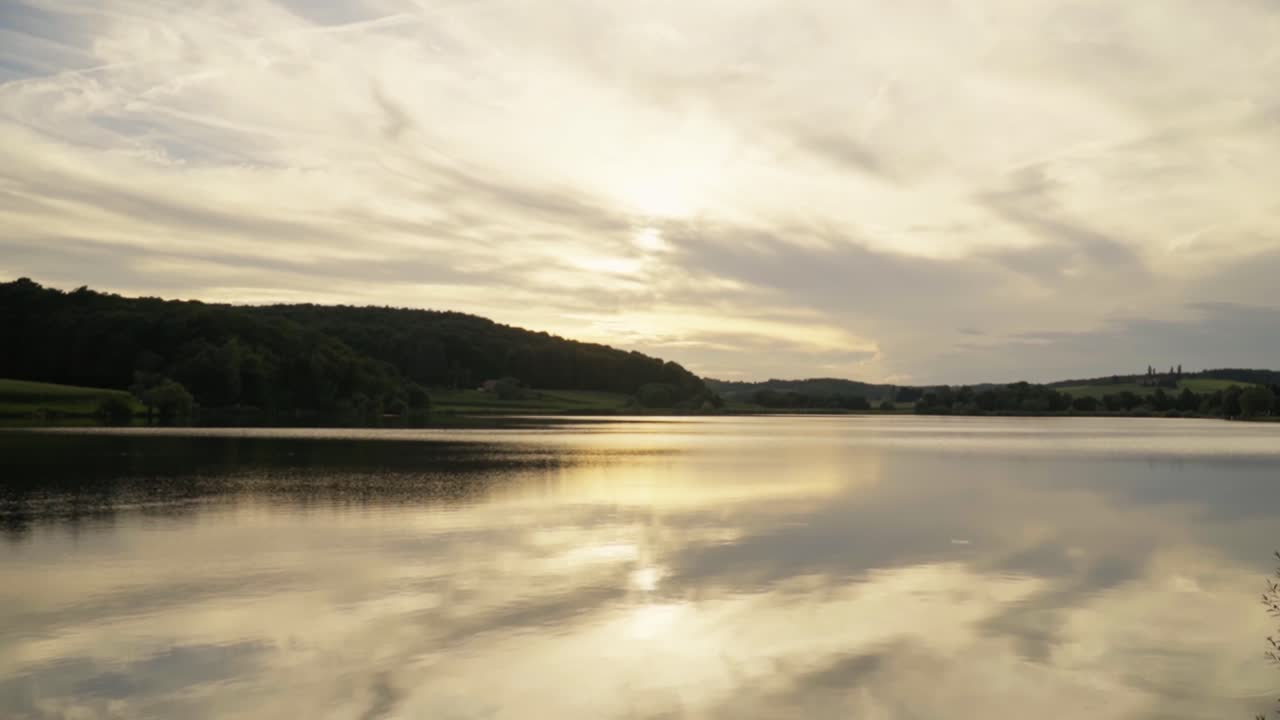 Wide view of calm lake reflecting golden sunset sky and soft clouds, capturing peaceful natural landscape and serene outdoor atmosphere in Savci Slovenia