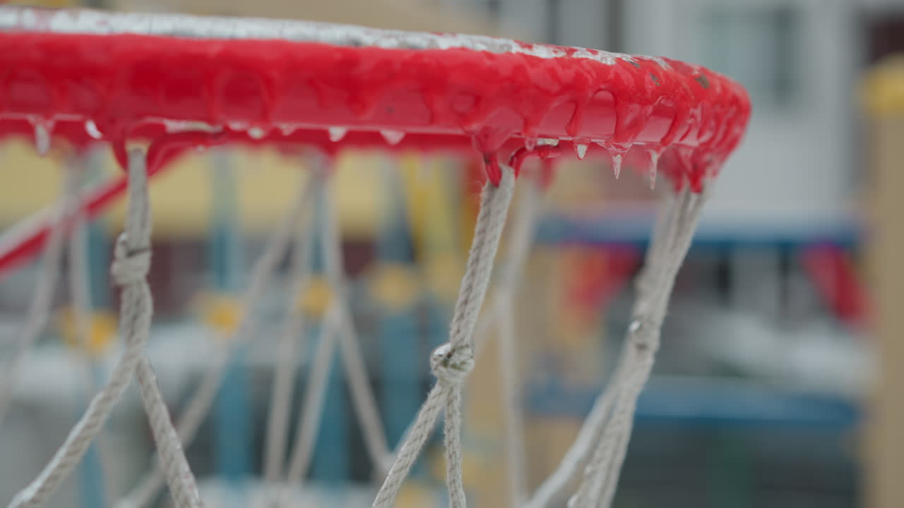 primer plano de un aro de baloncesto helado con hielos colgando del borde y un patio de recreo nevado borroso en el fondo, capturando la esencia del invierno y el clima frío durante la temporada