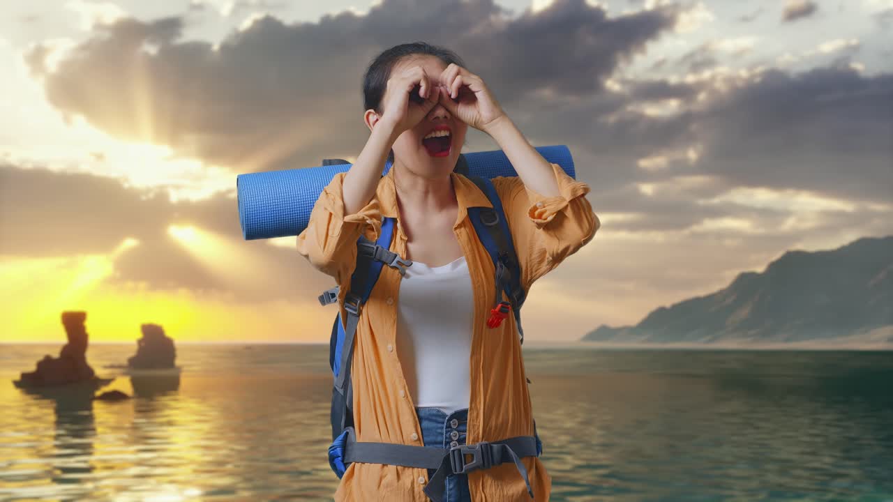 Mulher asiática praticante de hiking com mochila de montanhismo fazendo um gesto de binóculos, olhando para algo e dizendo "Uau" enquanto está em pé na praia durante o pôr do sol