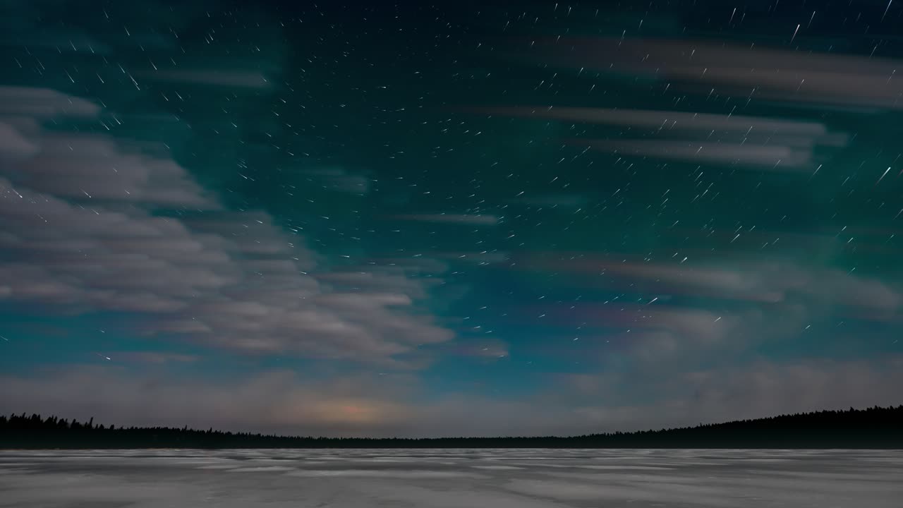 Timelapse looking at a turquoise colored night sky above a snow covered lake with clouds and stars moving and weak green colored Aurora.