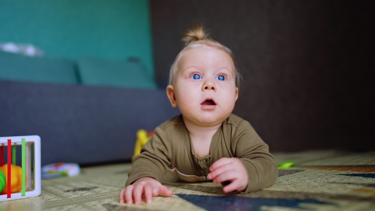 Criança loira pequena olhando surpresa com a boca aberta. Bebê adorável deitado no chão está focado em algo acima dele.