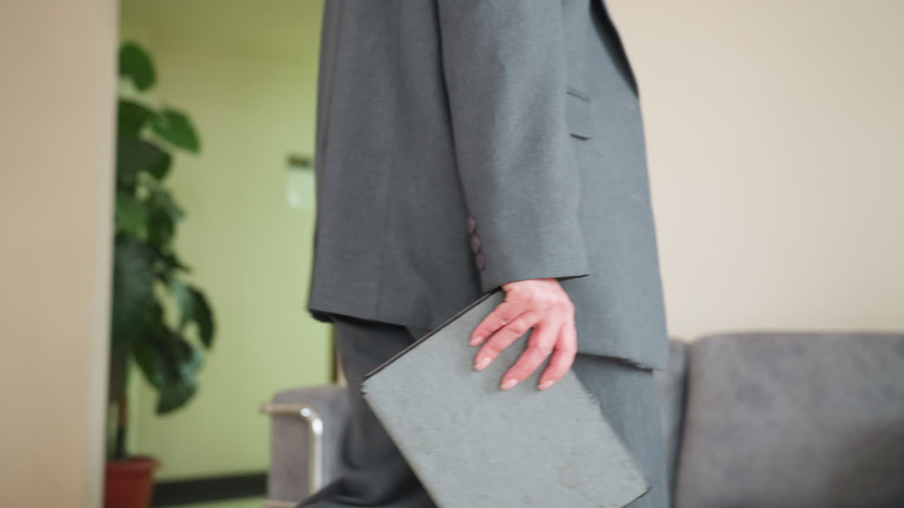 Close-up of business professional in gray suit holding document folder while walking toward office doorway. Corporate employee carrying paperwork through minimalist workplace