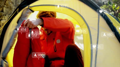 male camper reaching backpack inside yellow tent, displaying floating social media analytics icons