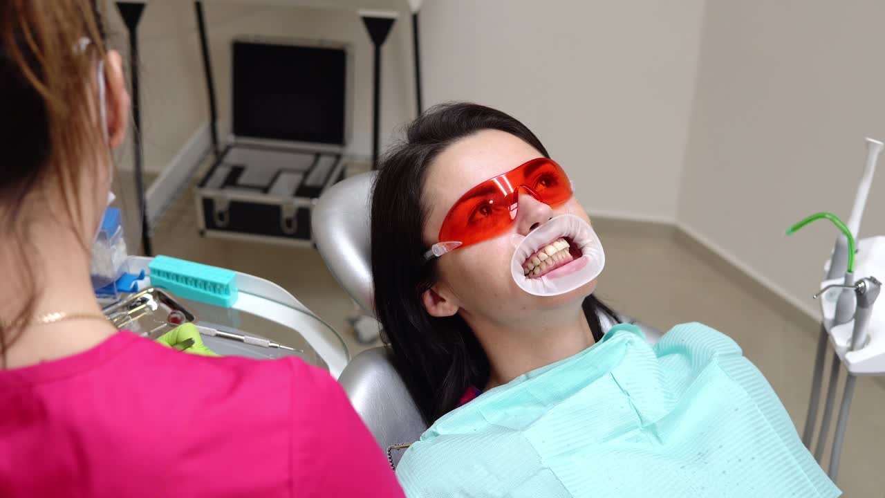 Young woman with an expander in mouth at the dental clinic. Application of whitening gel to the teeth. Modern dental office. Shot in 4k