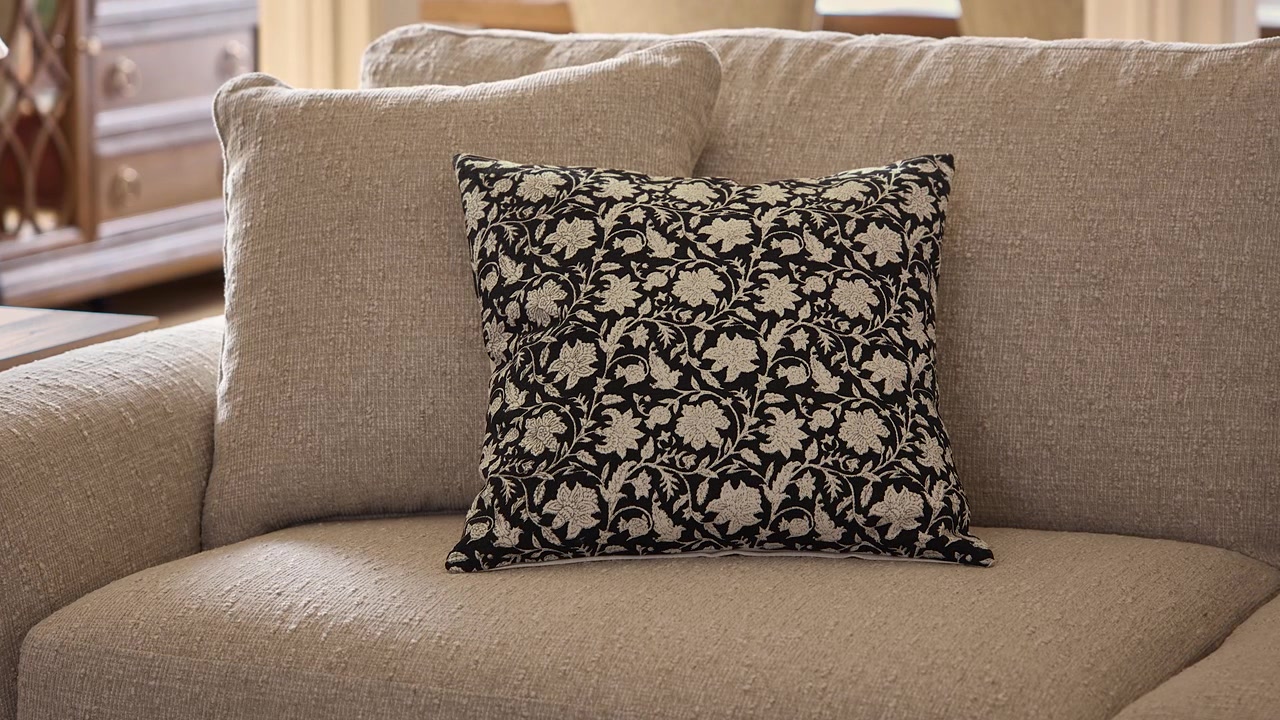 Långsam inzoomning på en svartvit blommig kudde som vilar på en grå soffa med varmt solljus som skiner över ett professionellt stylat rustikt vardagsrum